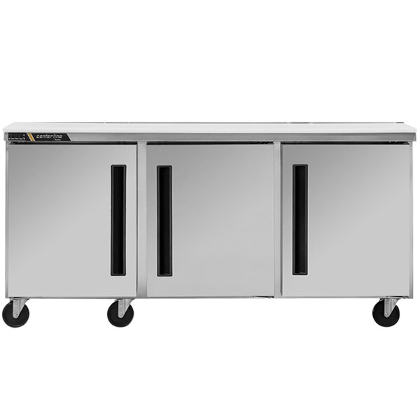 A silver Traulsen undercounter refrigerator with three left-hinged doors on wheels.
