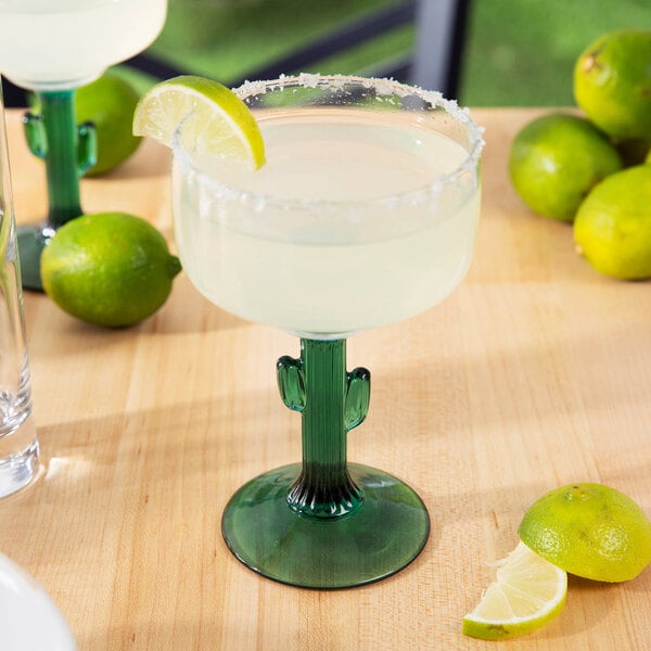 A Libbey cactus margarita glass with a drink on a table with lime wedges.
