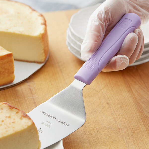 A hand using a Mercer Culinary purple pie server to cut a piece of cheesecake.