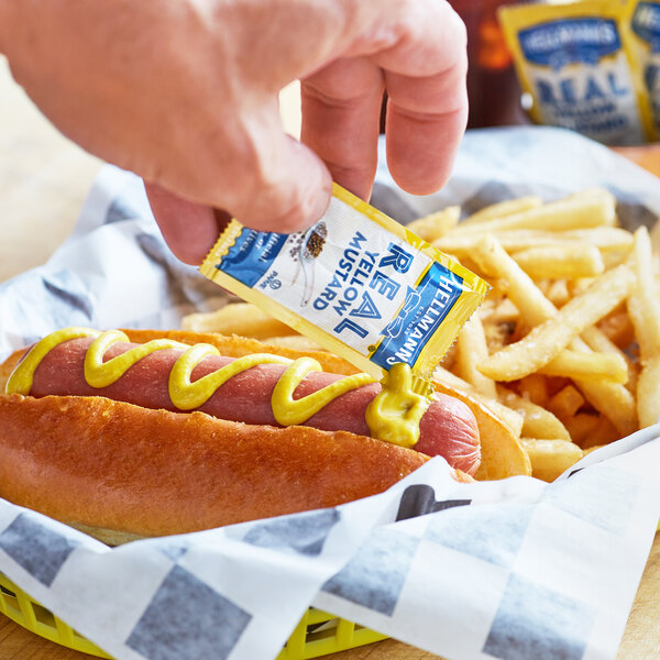 A person using a Hellmann's yellow mustard packet to put mustard on a hot dog.