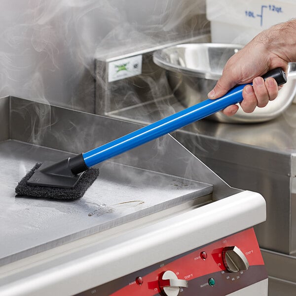 A hand with a blue Scrubble pad holder brush cleaning a stove.