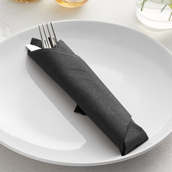 A fork and knife wrapped in a black Hoffmaster linen-like dinner napkin on a white plate.