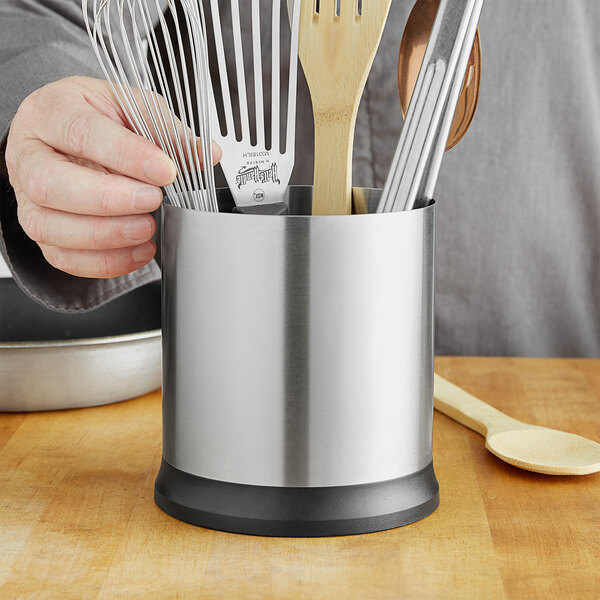 A person holding a silver and black OXO Good Grips rotating utensil holder filled with metal utensils.