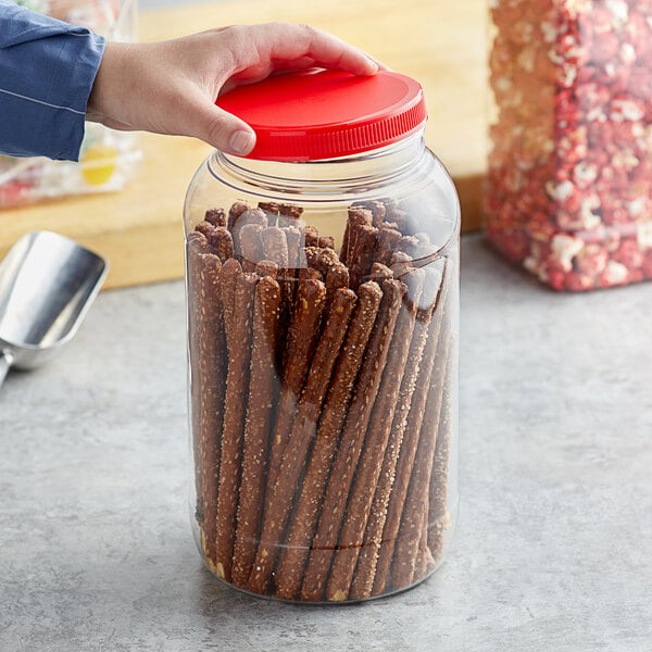 A hand holding a 1 gallon round PET plastic jar with a red lid filled with pretzels.
