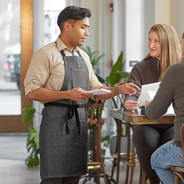A man in a Kennett gray denim Acopa bib apron serving food to a woman at a counter in a farm-to-table restaurant.