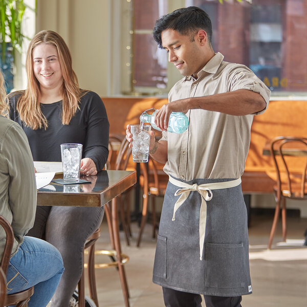 A man in an Acopa denim bistro apron serving water to a woman at a table.