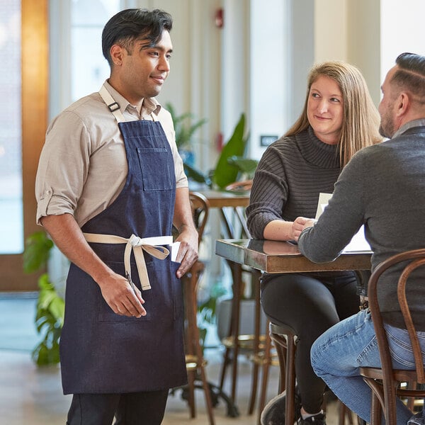 A man in a blue Acopa Kennett apron talking to a couple at a farm-to-table restaurant.