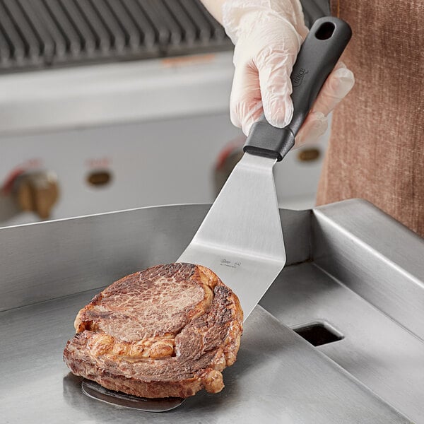 A hand holding a Choice perforated turner with a black handle over a steak.