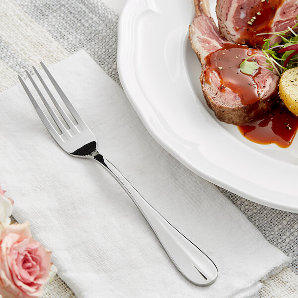 An Acopa stainless steel table fork on a napkin next to a plate of meat and vegetables.