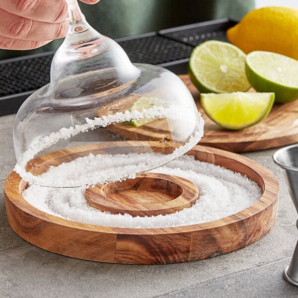 A hand using an Outset wood glass rimmer to salt a glass over a wooden dish.