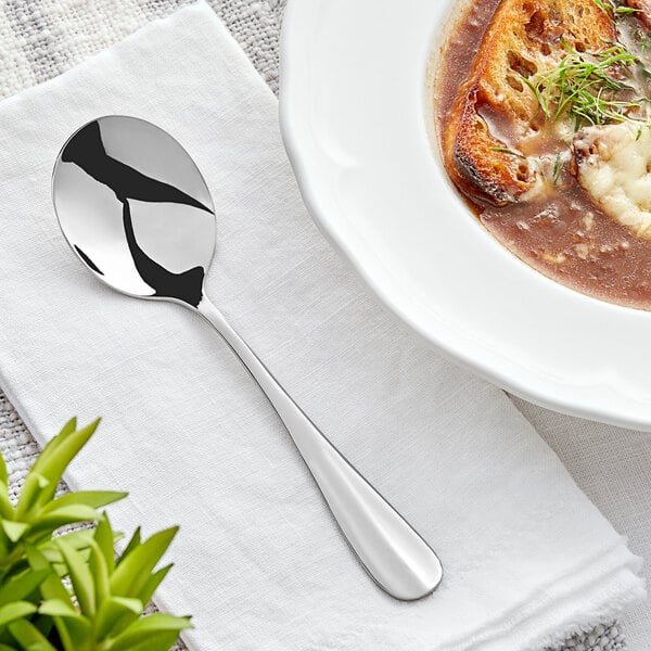 An Acopa stainless steel bouillon spoon on a bowl of soup on a white cloth.
