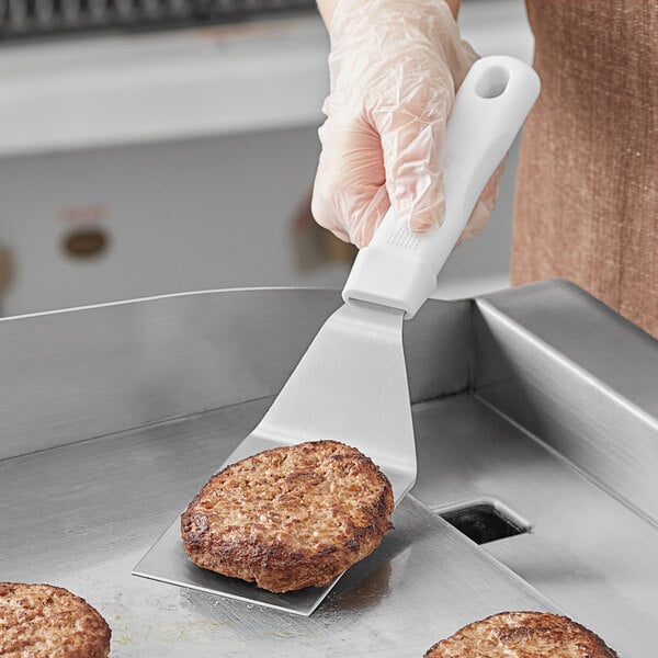 A person using a Choice beveled square edge turner to flip a burger patty on a grill.