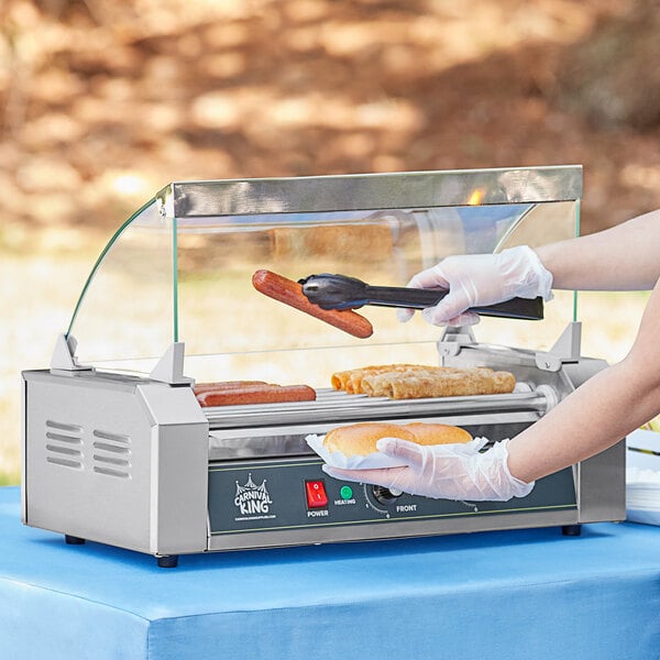 A person using a Carnival King Glass Sneeze Guard to serve hot dogs on a roller grill.