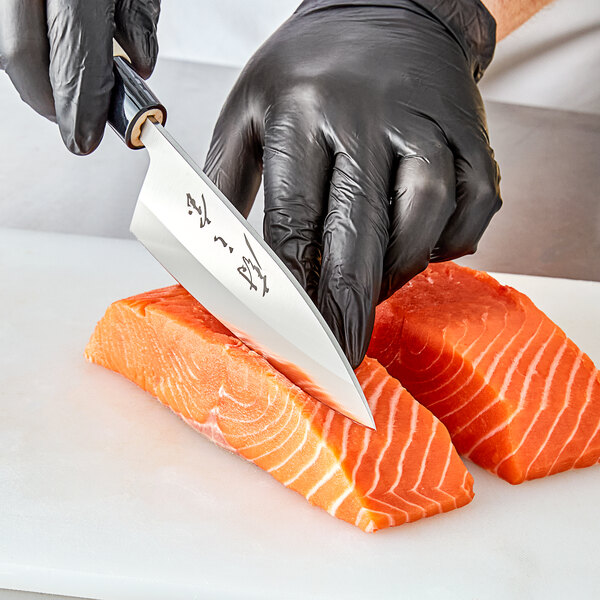 A person's gloved hand using a Mercer Culinary Deba knife to cut a piece of salmon.