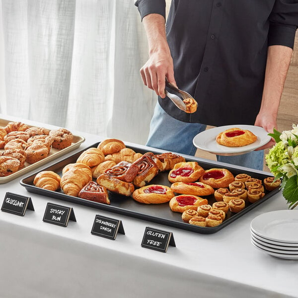 A person using a Baker's Mark black rimmed sheet pan to serve pastries on a buffet table.