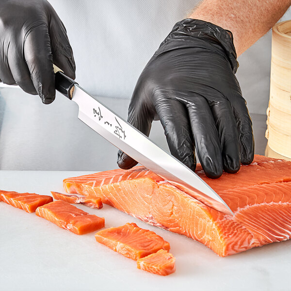 A person wearing black gloves using a Mercer Culinary sashimi knife to cut a piece of salmon.