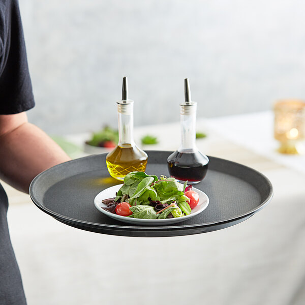 A person holding a Choice black round non-skid serving tray with salad and olive oil on it.