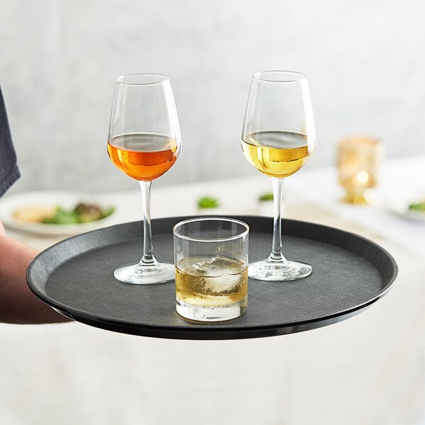 A person holding a Choice black non-skid serving tray with wine glasses and a glass of ice.