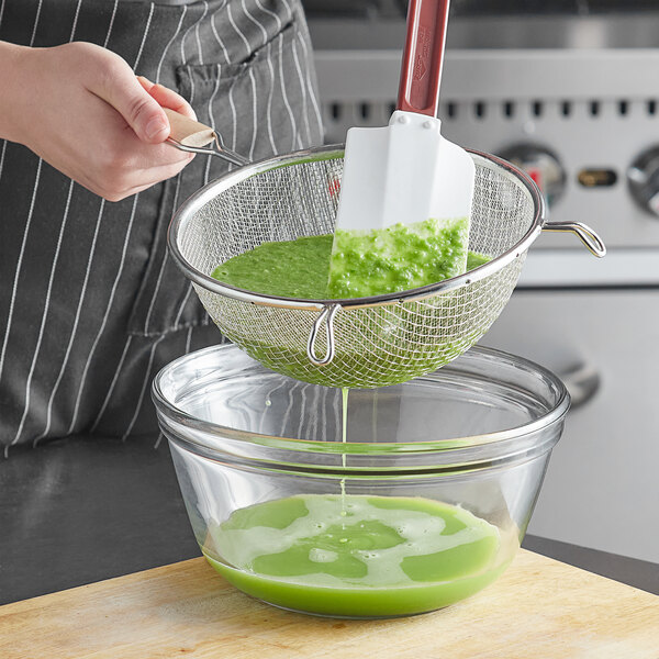 A person using a Vollrath medium mesh wire strainer with a wood handle to pour green liquid into a bowl.