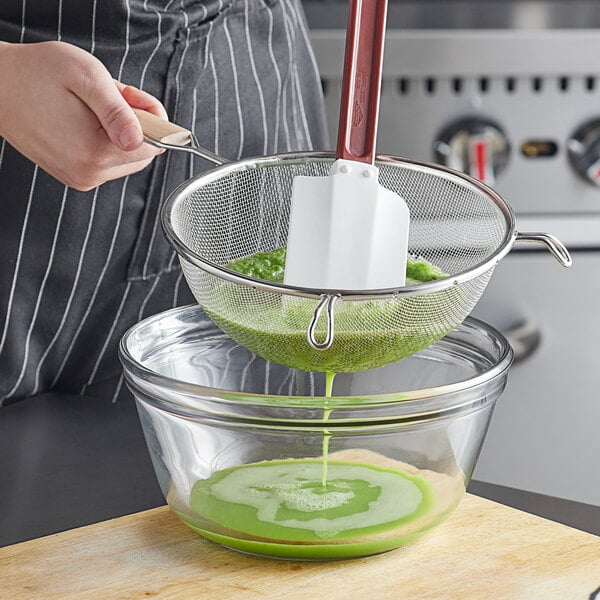 A person using a Vollrath medium mesh strainer with a wood handle to pour green liquid into a bowl.