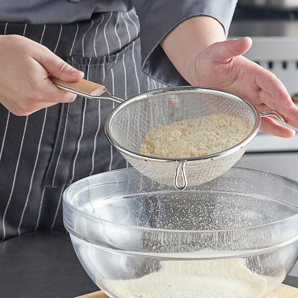 A person using a Vollrath double mesh strainer with a wooden handle to sift flour into a bowl.
