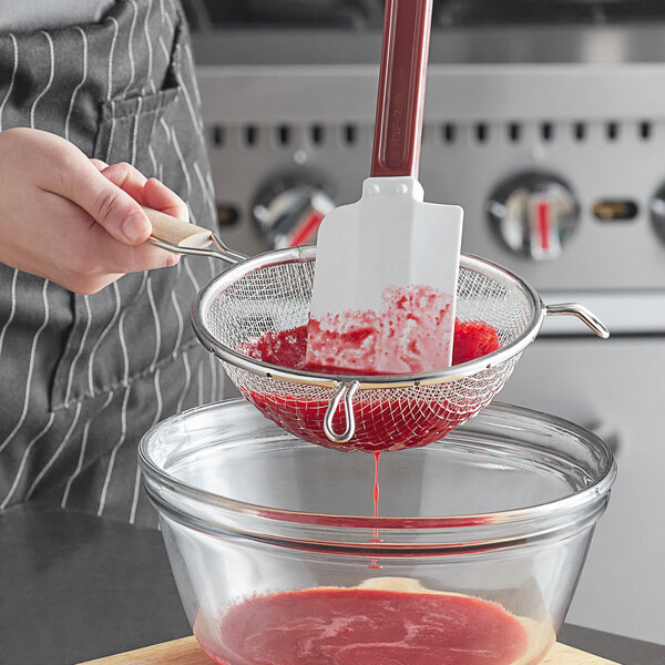 A person using a Vollrath medium mesh strainer with wood handle to strain red liquid.