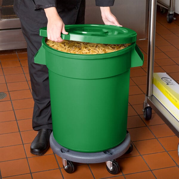 A woman in a chef's hat holding a green 20 gallon mobile ingredient storage bin full of pasta.
