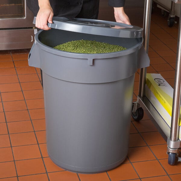 A woman opening a 32 gallon gray round ingredient storage bin with green food inside.