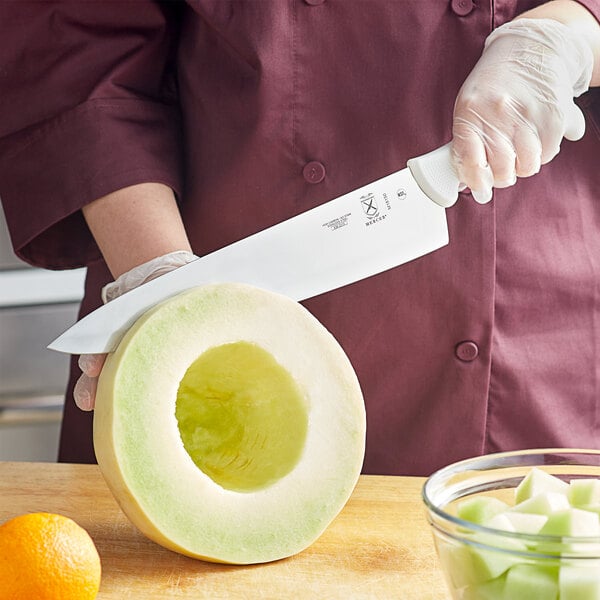 A hand holding a Mercer Culinary Ultimate White&#174; Chef Knife cutting a melon.