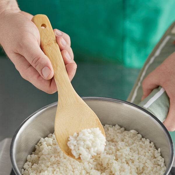 A hand holding Emperor's Select bamboo rice paddle stirring rice in a bowl.