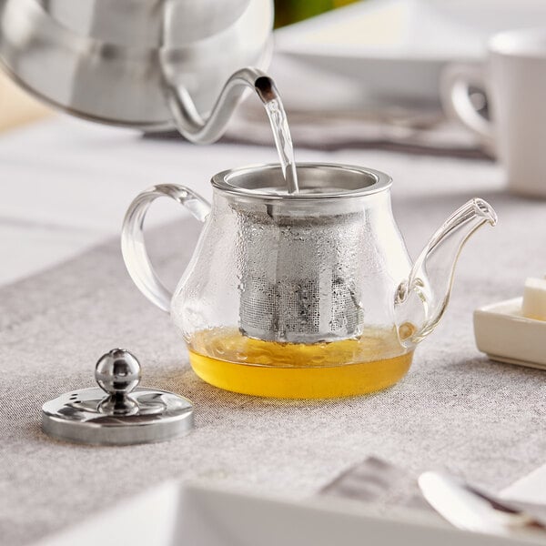 An Acopa glass teapot with a metal kettle pouring liquid into it.