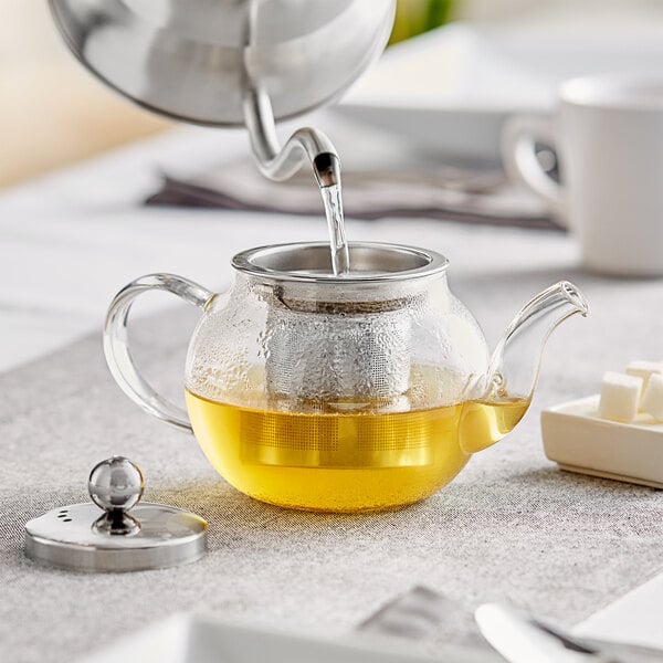 A glass teapot with a silver tea kettle pouring clear liquid into it.