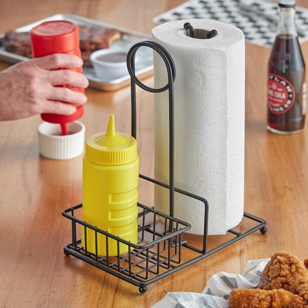 A Tablecraft black powder-coated metal paper towel holder holding paper towels and condiment bottles on a table.