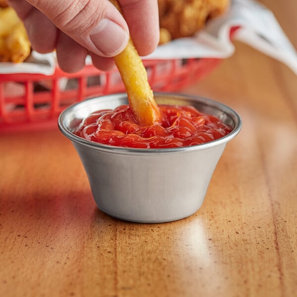 A person dipping a french fry into a Tablecraft stainless steel sauce cup filled with ketchup.