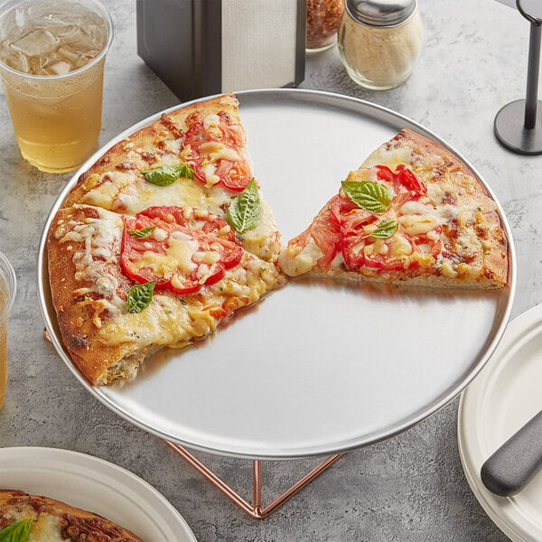 A Choice 13" aluminum coupe pizza pan with a slice of pizza topped with tomatoes and basil on a white plate.