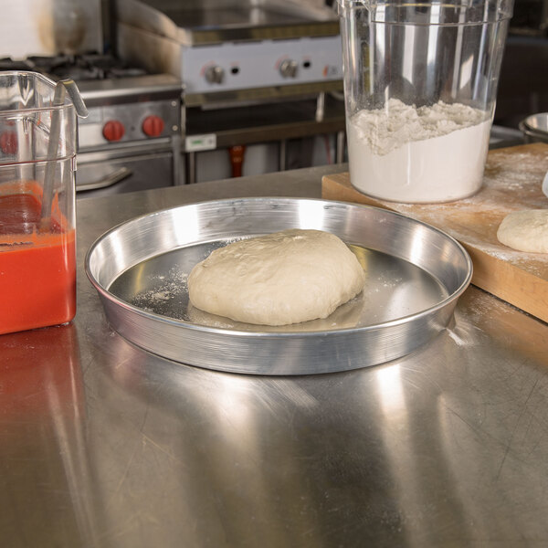 An American Metalcraft aluminum pizza pan with dough on it on a counter next to a container of flour.