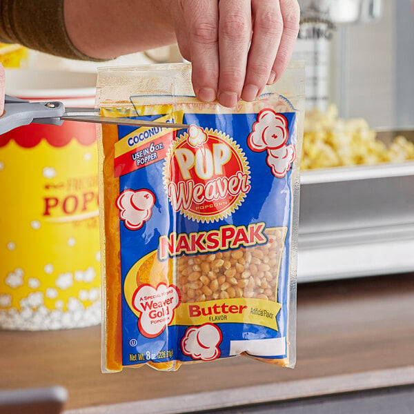 A person holding a bag of Pop Weaver All-In-One Naks Pak popcorn in front of a popcorn machine.