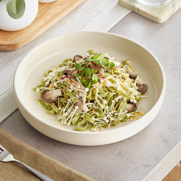 An Acopa Pangea fog white porcelain pasta bowl filled with pasta, mushrooms, and sprouts on a table with a brown cloth and a silver spoon.
