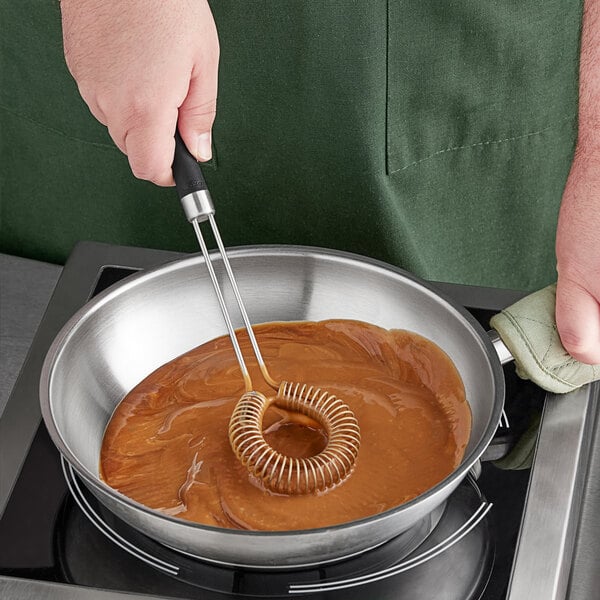 A person stirring brown liquid in a pan with an OXO Good Grips metal sauce whisk.