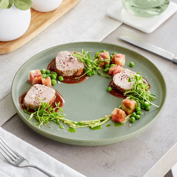 An Acopa Pangea sage matte porcelain plate with meat, peas, and a white object on it on a table with a fork and knife.