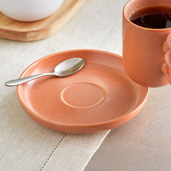 A hand holding an Acopa Terra Cotta saucer with a cup of coffee and a spoon.