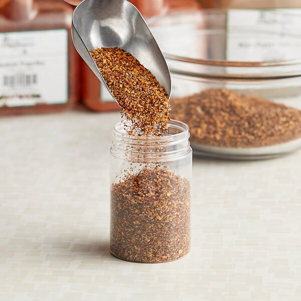 A metal scoop pouring brown seasoning into a clear plastic jar.