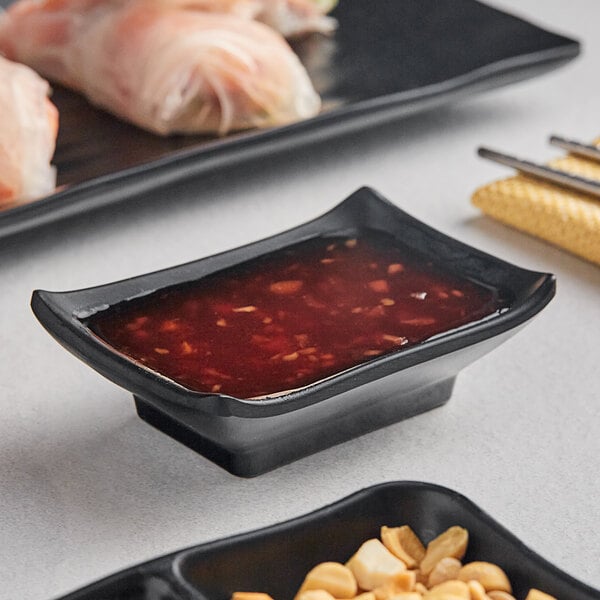 A black rectangular Acopa Izumi sauce dish with red sauce in it next to a plate of food.