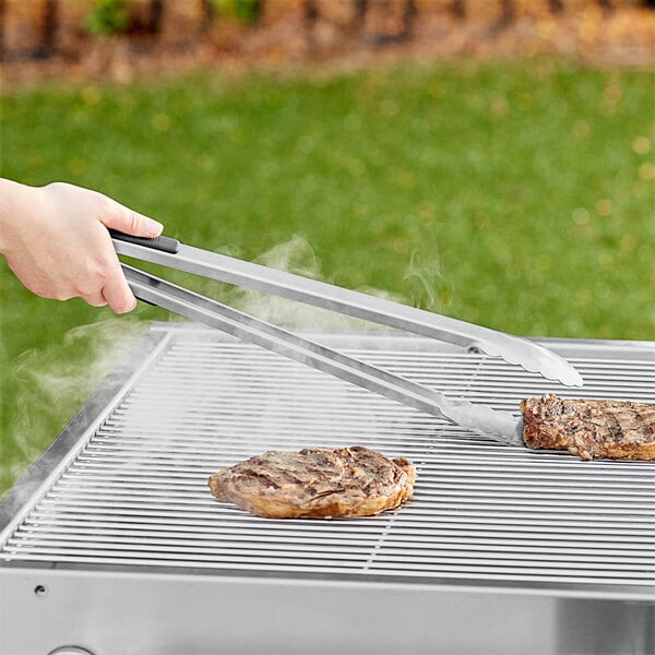 A hand using Backyard Pro stainless steel tongs to grill meat over smoke.