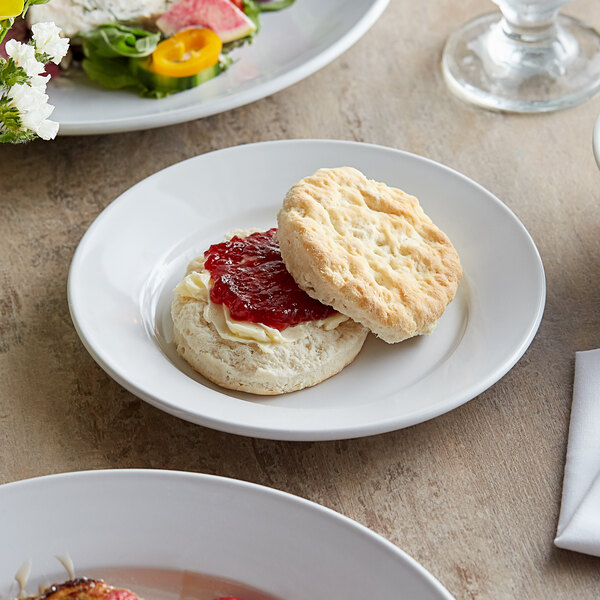 A biscuit with jam on a white Acopa stoneware plate next to a glass of tea.