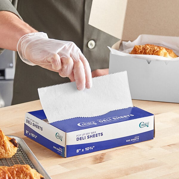A person wearing gloves holding Choice interfolded wax paper over a box of food.