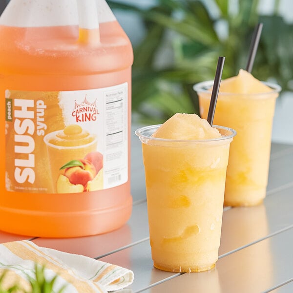 A plastic cup with a yellow peach slushy drink in it next to a bottle of Carnival King Peach Slushy Concentrate.