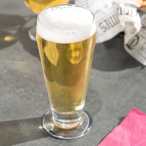 A Libbey footed pilsner glass full of beer with foam on a table.