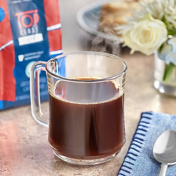 A glass mug of Lavazza coffee with a spoon and a bag of Lavazza Top Class Filtro Coarse Ground Coffee on a table.