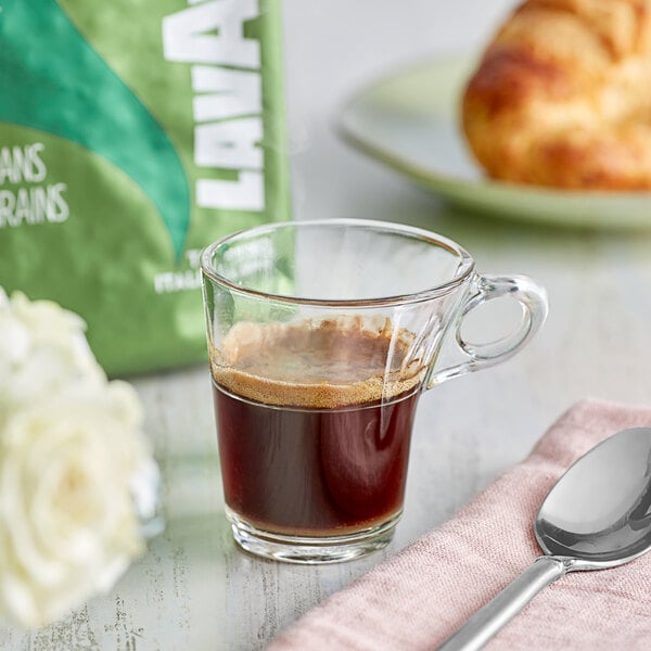 A glass cup of Lavazza Tierra! Brasile Intense Espresso on a table with a spoon and bag of whole bean coffee.
